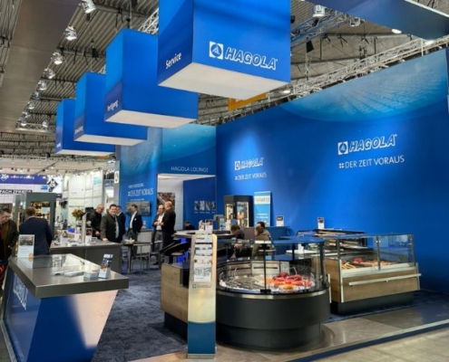 Auf diesem Foto sehen wir den Messeauftritt von HAGOLA. Das Motto auf der Gastroleitmesse des deutschen Herstellers von hochwertigen Schanktischen usw. lautet - Hagola - der Zeit voraus....
